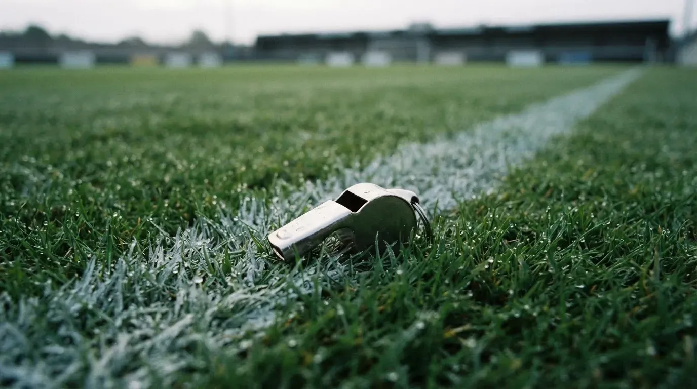 Eine Trillerpfeife liegt auf dem Rasen neben der Mittellinie, Tau hängt an den Halmen, im Hintergrund ein leerer Spielfeldrand.