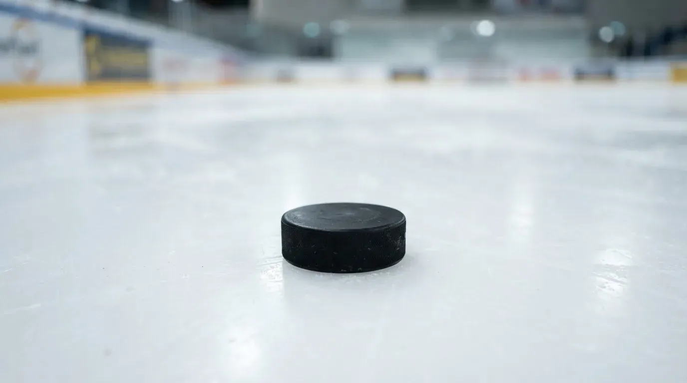 Ein Eishockeypuck liegt auf dem Eis, im Hintergrund unscharf die Bande unter hellem Hallenlicht.