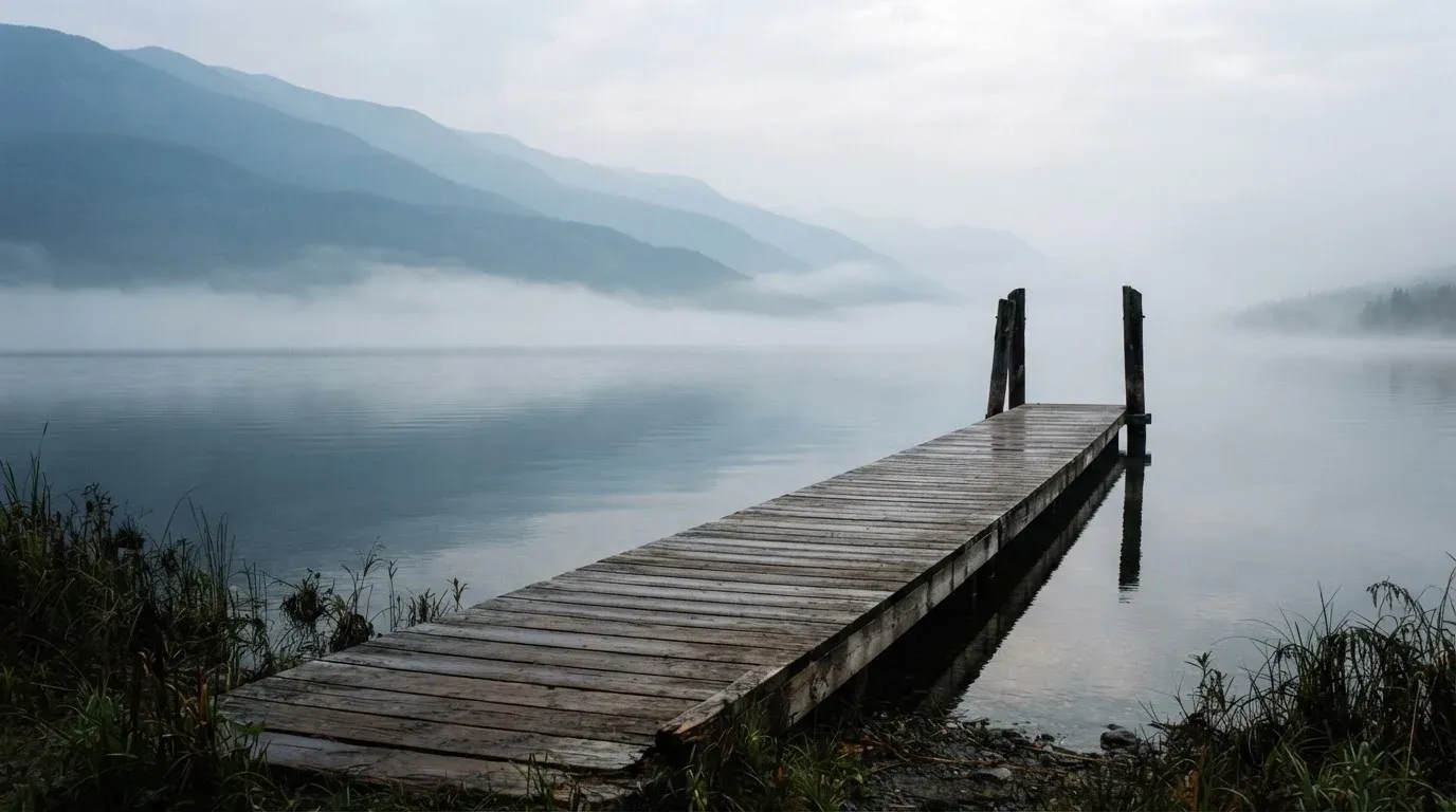 Ein Seeufer mit einem hölzernen Steg, Nebel liegt über dem Wasser, am anderen Ufer erheben sich Berge.