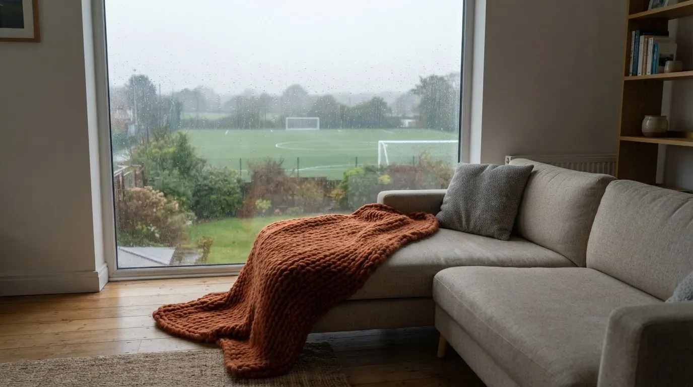 Ein Sofa mit Wolldecke steht vor einem grossen Fenster, draussen Regen und in der Ferne ein Fussballplatz.