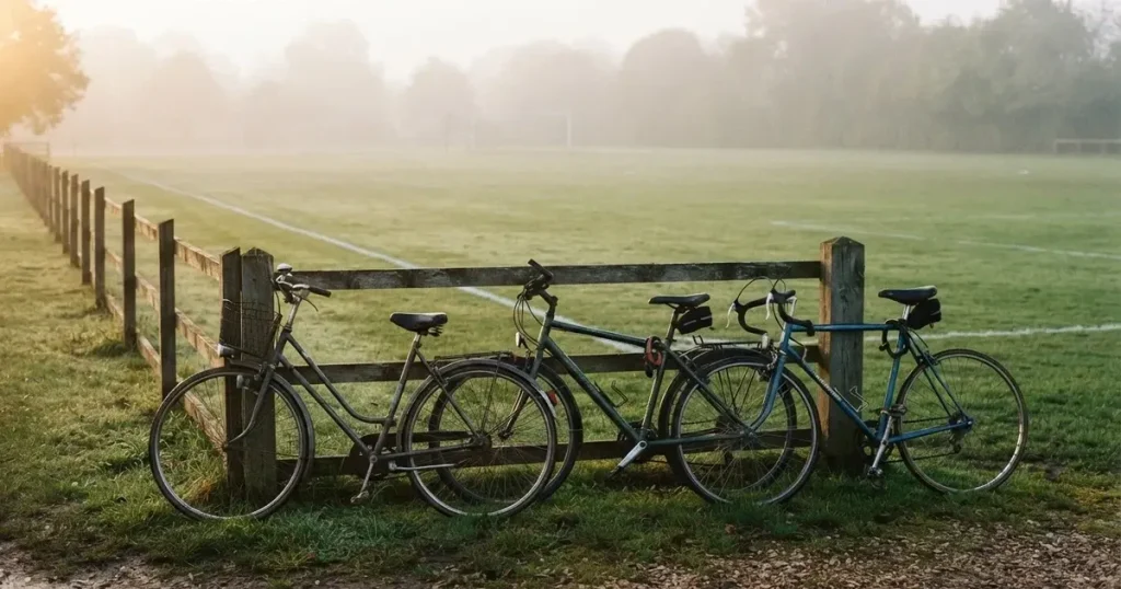 Drei Fahrräder lehnen nebeneinander an einem Zaun vor einem Sportplatz, dahinter ein grüner Rasen im Dunst.