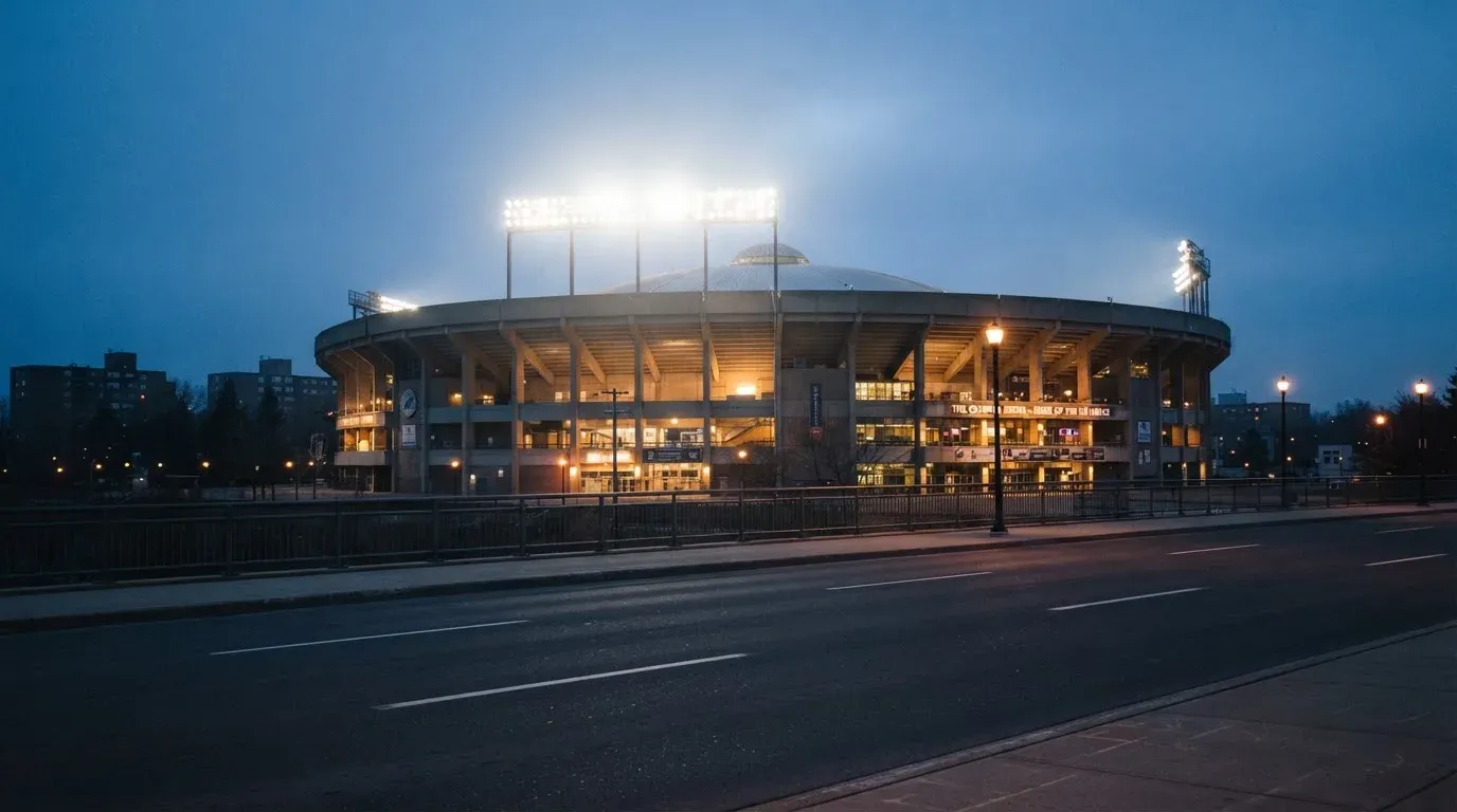 Ein grosses Eisstadion aus der Ferne gesehen, hell erleuchtet von Flutlicht in der Abenddämmerung, davor eine leere Strasse.