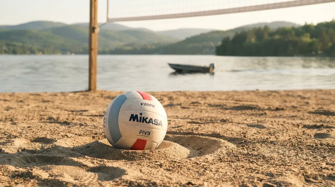 Ein Volleyball liegt im Sand eines Spielfelds am Seeufer, im Hintergrund gleitet ein Boot über das Wasser.
