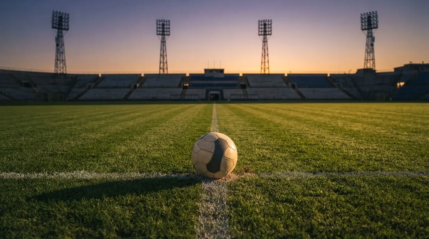 Ein Fussball liegt auf dem Mittelkreis eines leeren Rasenplatzes, dahinter ragen leere Tribünenreihen in den Abendhimmel.