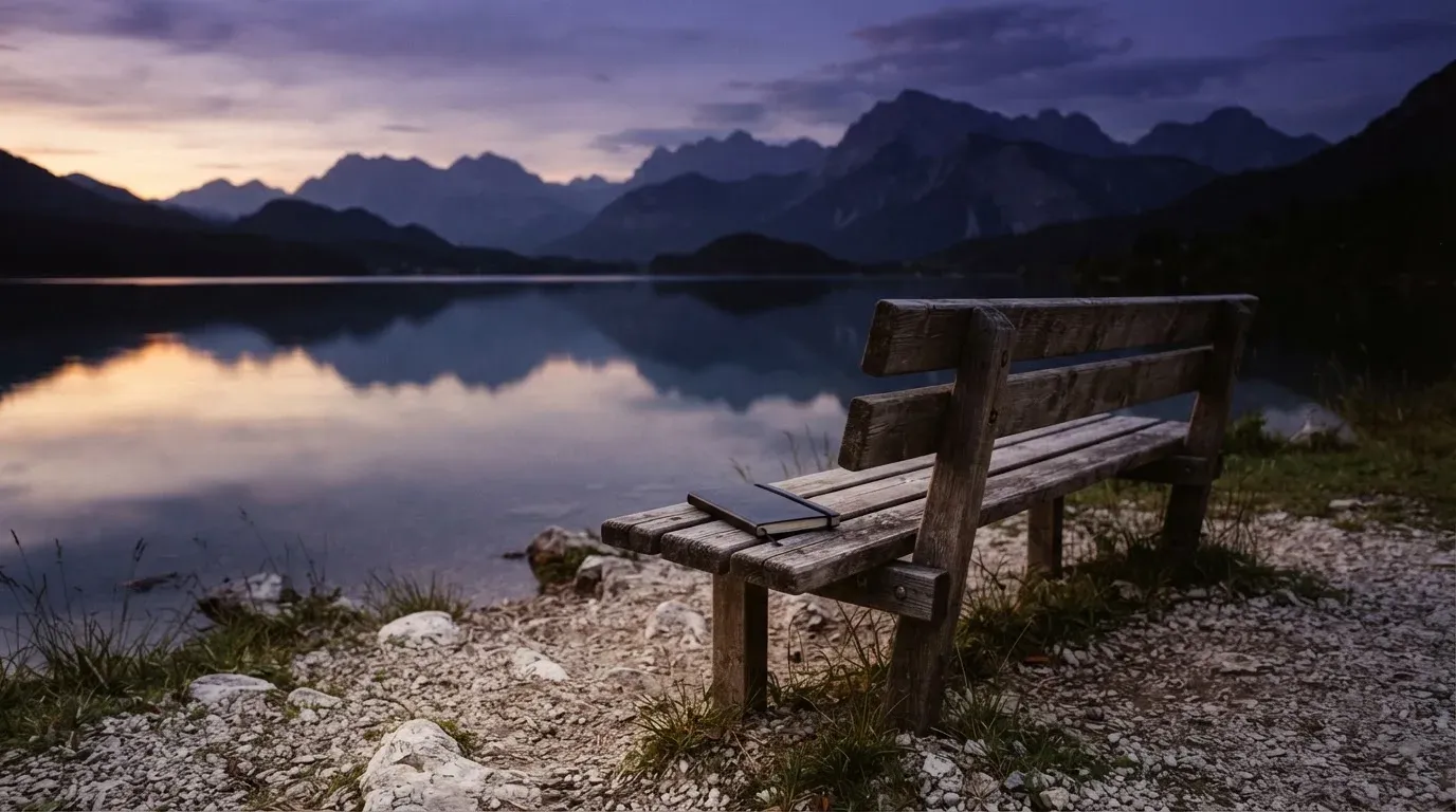 Eine Holzbank steht in der Dämmerung am Seeufer, im Hintergrund zeichnet sich ein Bergpanorama gegen den violetten Himmel ab.