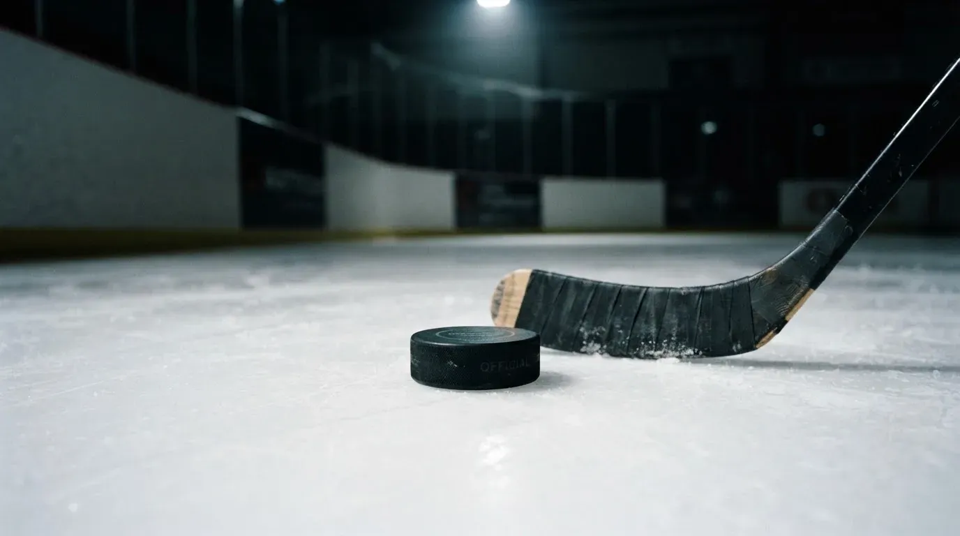 Ein Eishockeypuck liegt auf der Eisfläche neben dem Blatt eines Schlägers, die Bande dahinter ist im Dunkeln nur schwach zu erkennen.