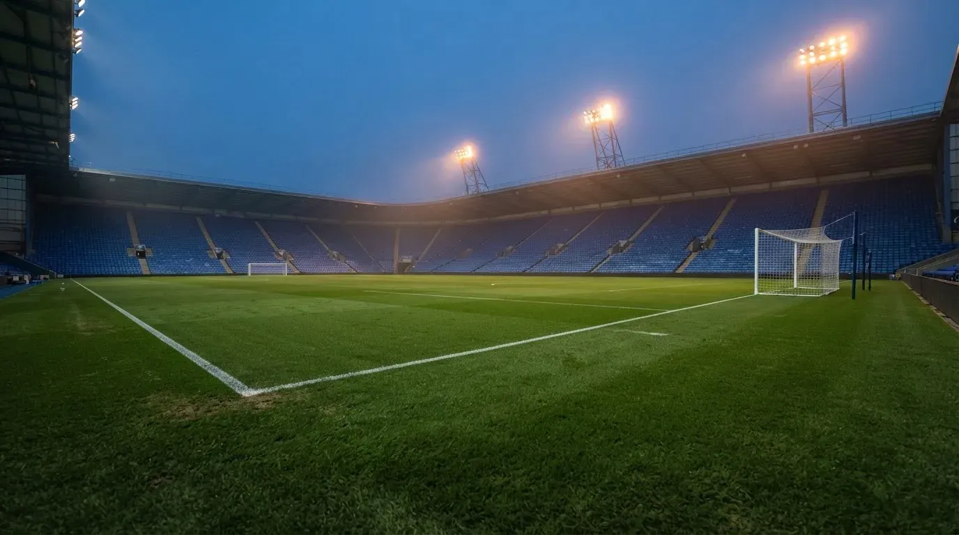 Leere Tribünenränge eines Fussballstadions in der Abenddämmerung, über dem Rasen leuchten die Flutlichter.