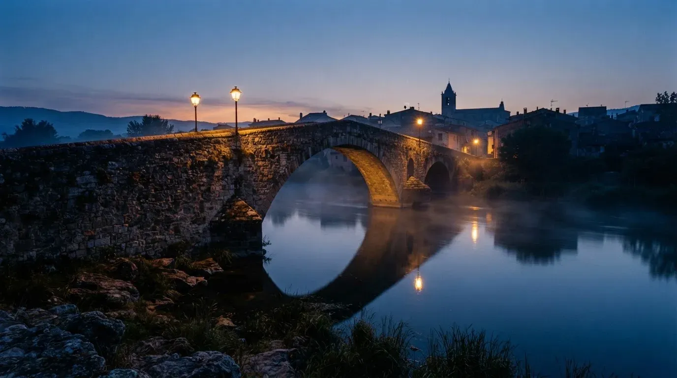 Eine Steinbrücke spannt sich in der Dämmerung über einen Fluss, auf dem Wasser liegt leichter Nebel.