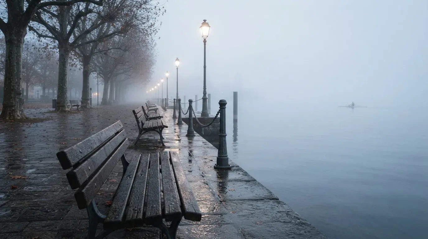 Eine Seepromenade mit leeren Bänken an einem nebligen Morgen, im Wasser spiegeln sich die Lichter der Uferlaternen.
