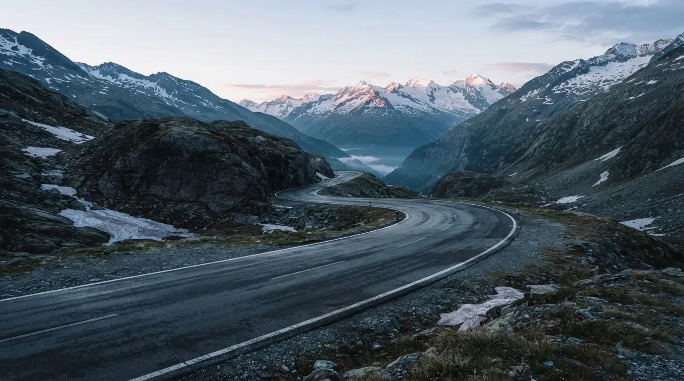 Ein Bergpass in den Schweizer Alpen mit leerer Strasse und Schneeresten am Rand im ersten Frühlicht.