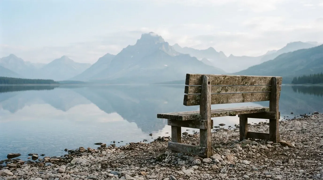 Eine Holzbank an einem Seeufer mit Blick auf eine Bergkette im Morgendunst, das Wasser ist spiegelglatt.