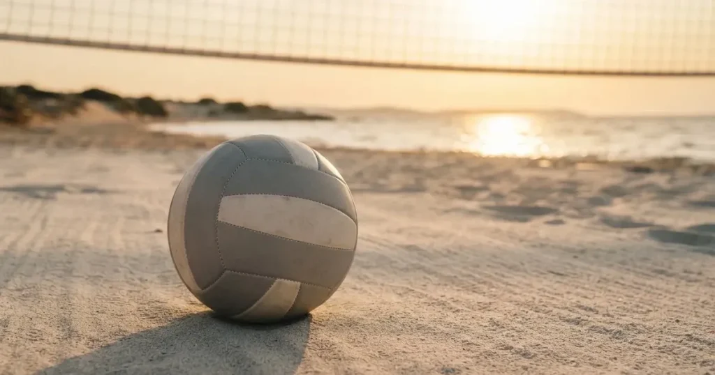 Ein Volleyball liegt auf feinem hellem Sand, ein gespanntes Netz verschwimmt dahinter, das Licht kommt von einem weiten Meer.