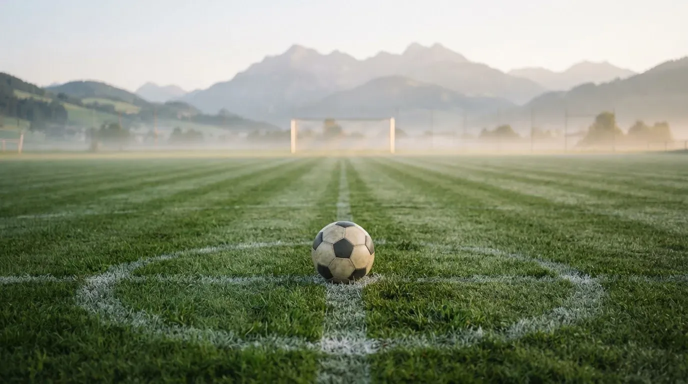 Ein einzelner Fussball auf dem Mittelpunkt eines leeren Rasens im Morgennebel, hinter dem Tor ragen Berge auf