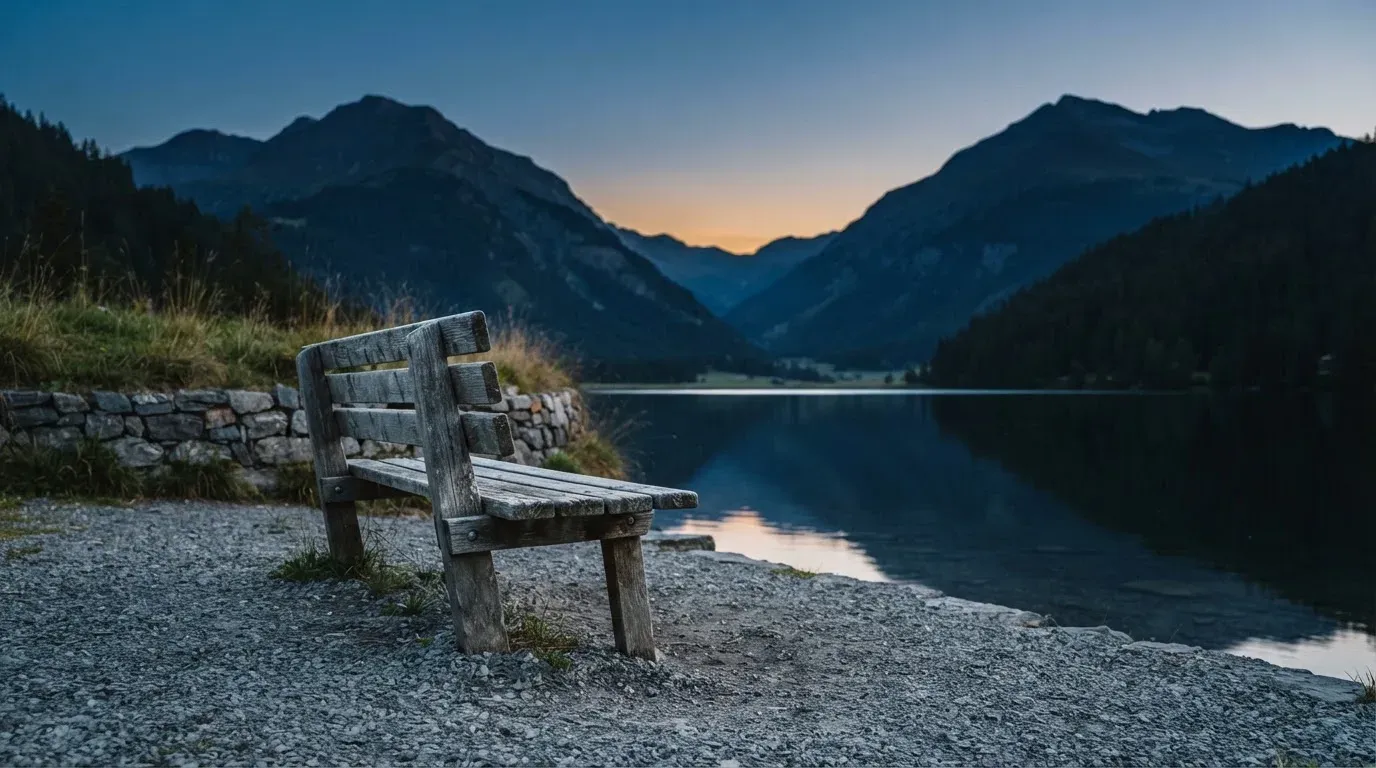 Eine leere Holzbank am Seeufer in der Dämmerung, im Hintergrund dunkle Berge