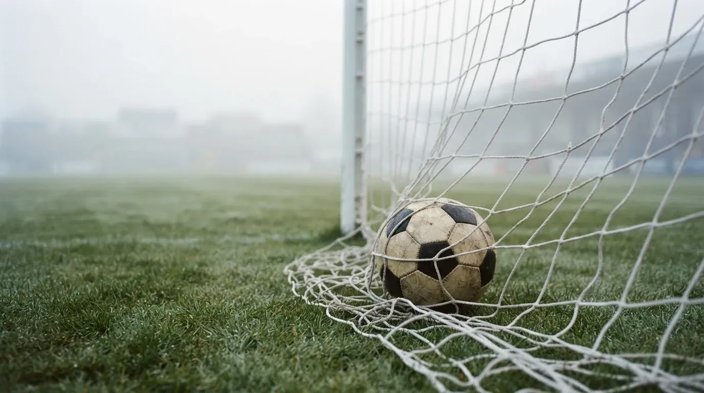 Ein Fussball liegt im Netz hinter einem Tor, weicher Morgennebel hängt über dem Rasen