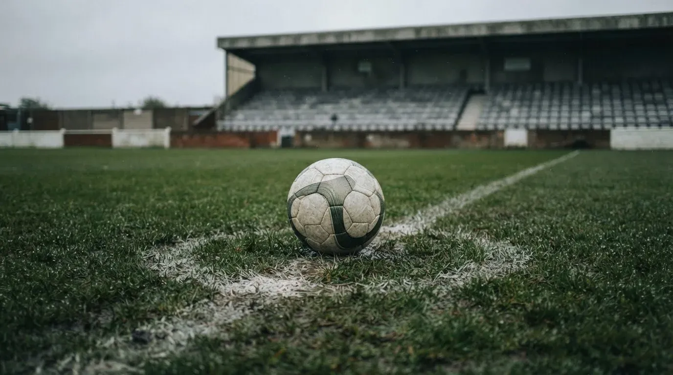 Ein Fussball liegt auf dem Anstosspunkt eines nassen Rasens, dahinter eine leere Tribüne unter grauem Himmel.