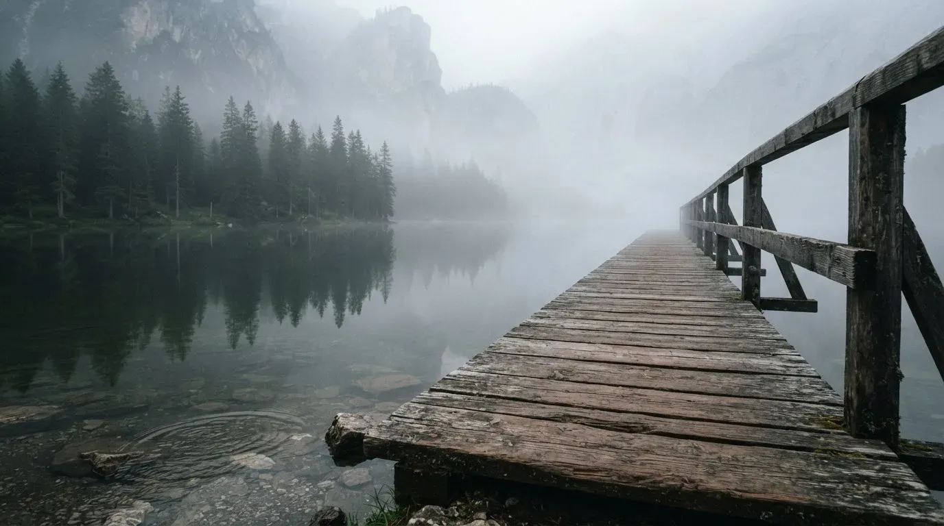 Ein Holzsteg führt über einen ruhigen Bergsee im Nebel, am anderen Ufer stehen dunkle Tannen.