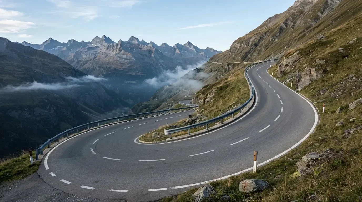 Eine Passstrasse in den Alpen mit enger Kurve und Leitplanke, darüber ein klarer Morgenhimmel und einzelne Nebelschwaden im Tal.