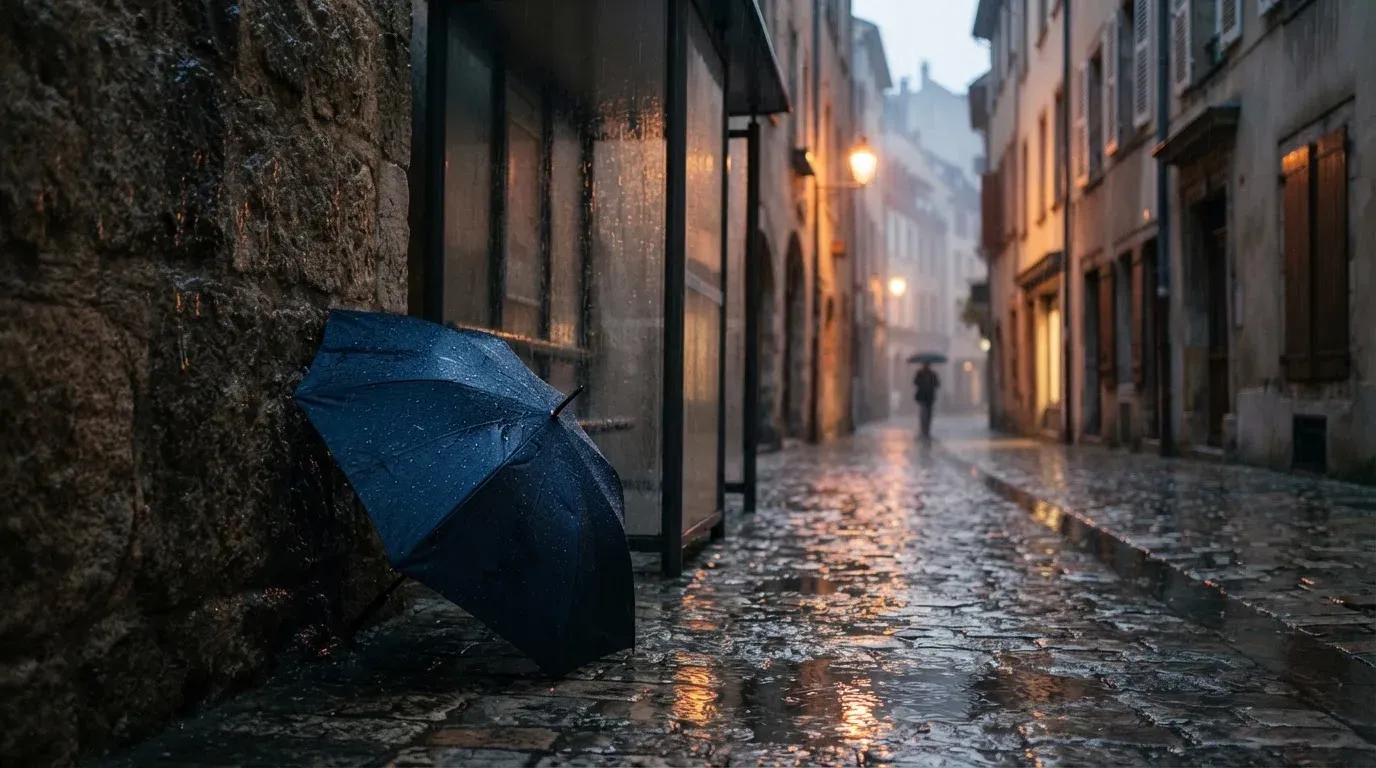 Eine Tramhaltestelle bei Regen in einer Altstadtgasse, das Kopfsteinpflaster glänzt und ein einzelner Regenschirm lehnt an einem Mäuerchen.