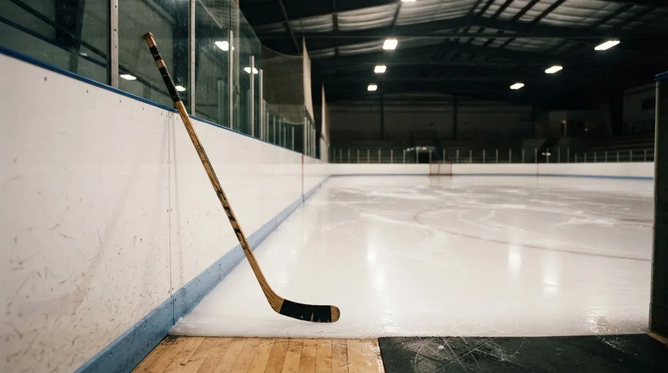 Ein Eishockeyschläger lehnt an der Bande, in der Ferne liegt die hell beleuchtete Eisfläche einer Halle unter Flutlicht.