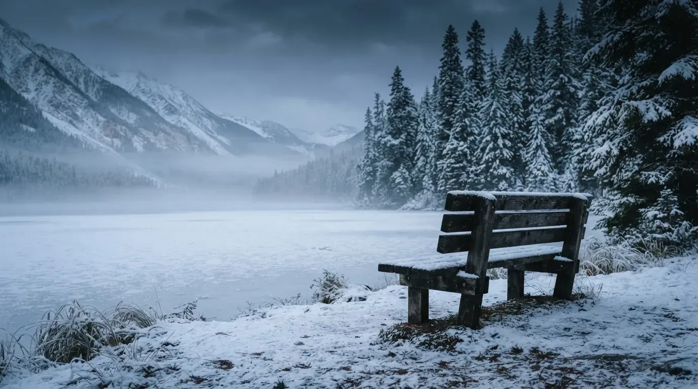 Ein zugefrorener Bergsee im Morgendunst, am Ufer steht eine einsame Holzbank zwischen verschneiten Tannen.