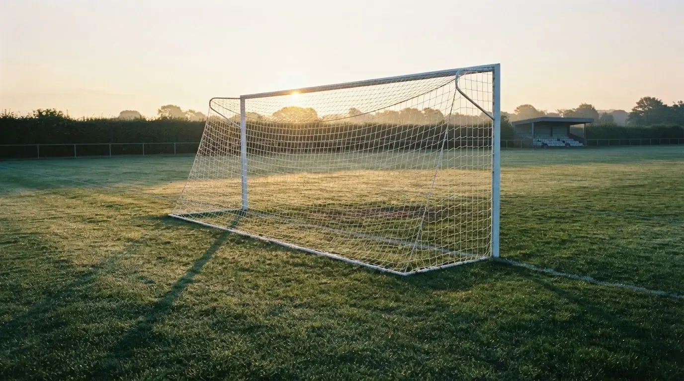 Ein Fussballtor mit Netz in der Totalen auf einem leeren Platz, Tau glänzt im Gras und die Morgensonne steht tief.