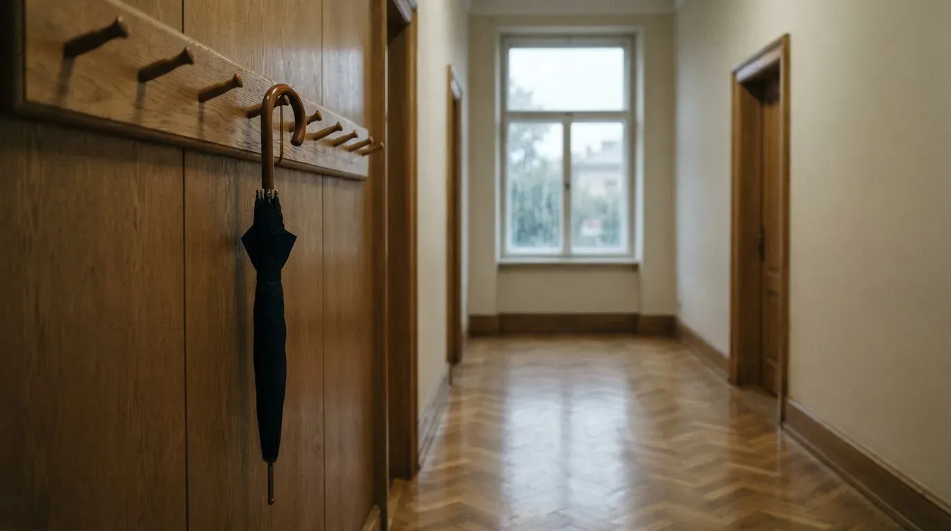 Eine Garderobe aus Holz mit einem dunklen Regenschirm in einem stillen Korridor, am Fenster rinnt Regen herunter.