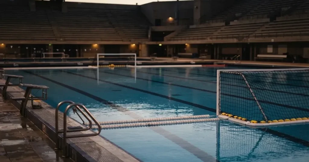 Ein Wasserballbecken am Abend mit zwei schwimmenden Torrahmen an den Enden, ruhiges blaues Wasser, leere Ränge.
