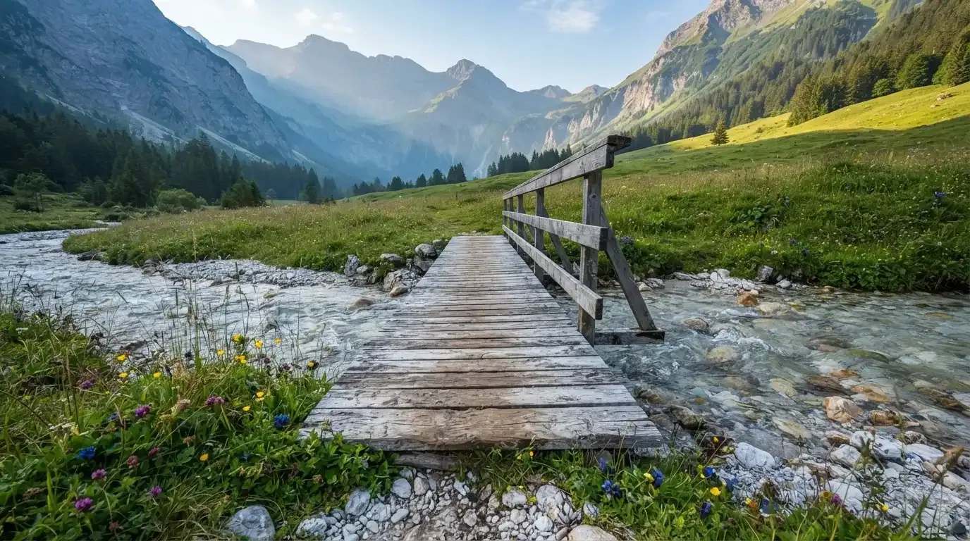 Ein Holzsteg führt über einen klaren Bergbach, im Hintergrund Wiesen und steile Hänge im Morgenlicht.