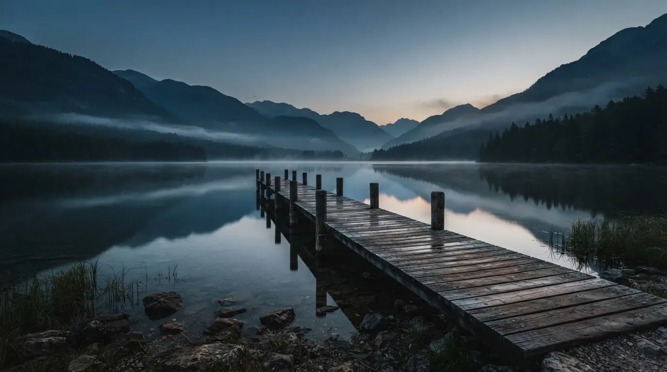 Ein Bergsee in der Dämmerung mit einem leeren Holzsteg, die Wasseroberfläche ist ruhig und Nebel zieht über die Hänge.