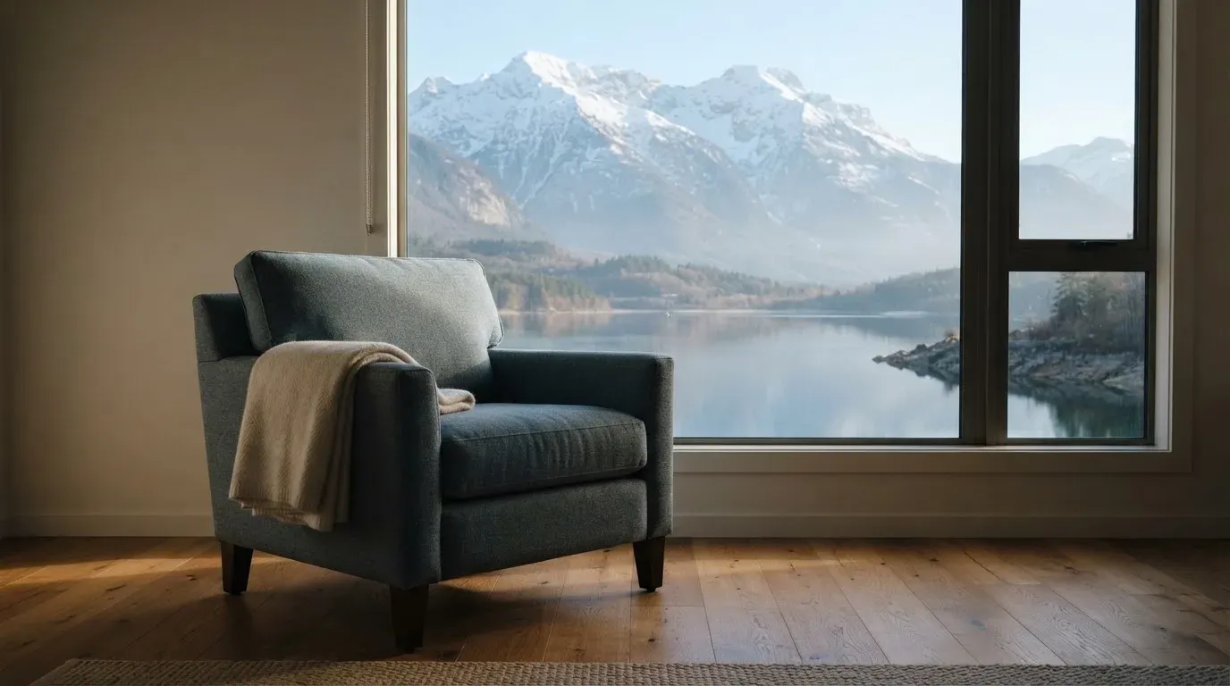 Ein leerer Sessel steht an einem Fenster mit Blick auf einen ruhigen See und schneebedeckte Berge im Morgenlicht.