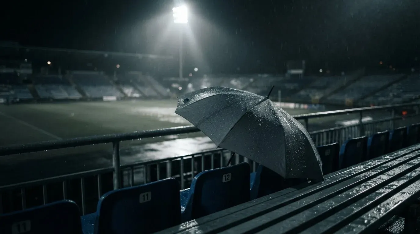 Ein aufgespannter Regenschirm liegt auf einer leeren Tribünenbank, Regentropfen glitzern im Licht der Flutlichter.
