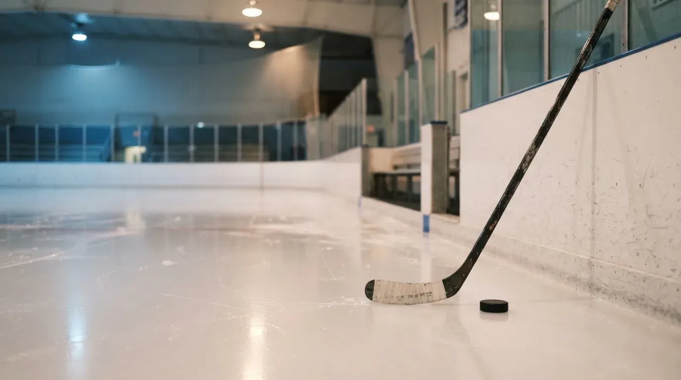 Ein Eishockeyschläger lehnt an der Bande einer leeren Halle, auf dem Eis spiegeln sich die Deckenlampen.