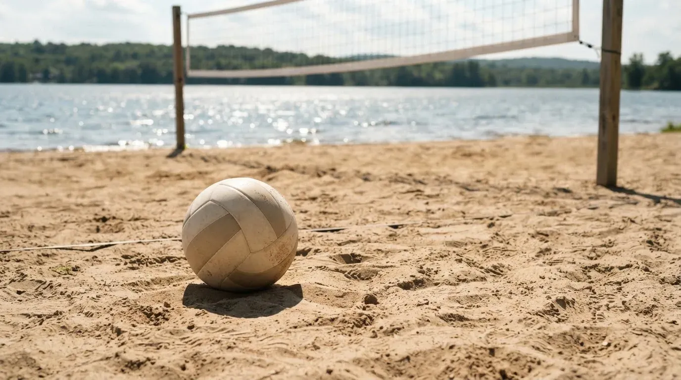 Ein Volleyball liegt auf dem Sand eines Strandfeldes am Seeufer, im Hintergrund glitzert das Wasser.