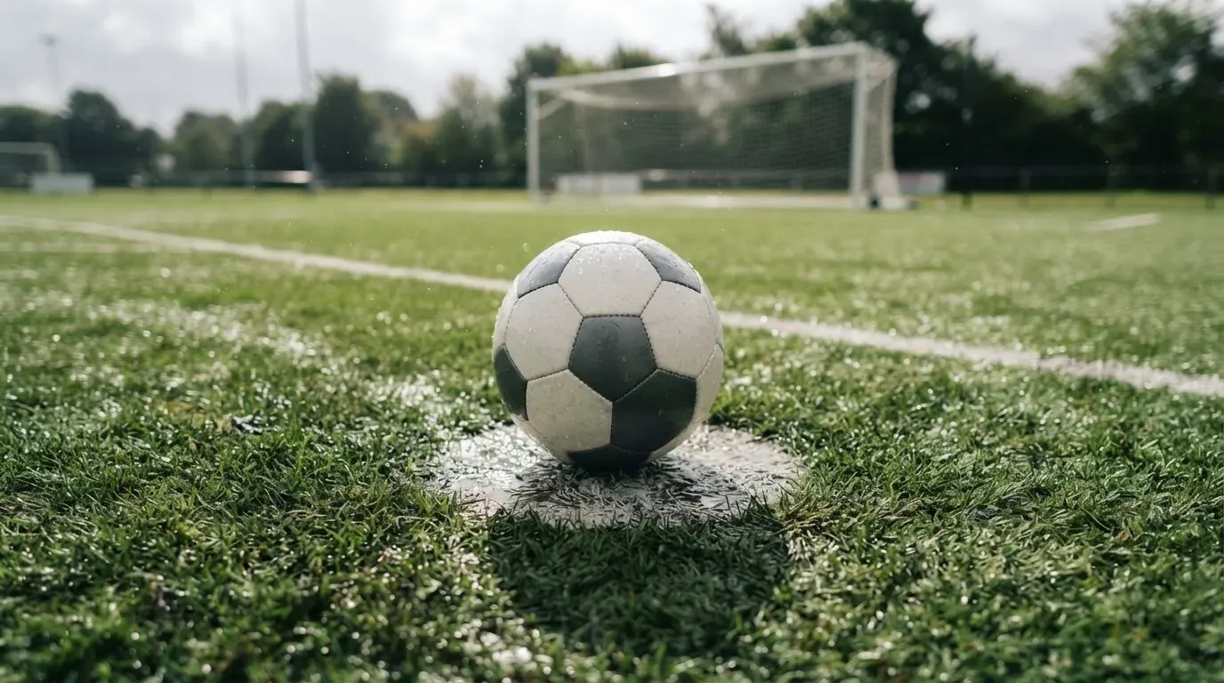 Ein einzelner Fussball liegt auf dem Elfmeterpunkt eines leeren Strafraums, der Rasen glänzt nass.