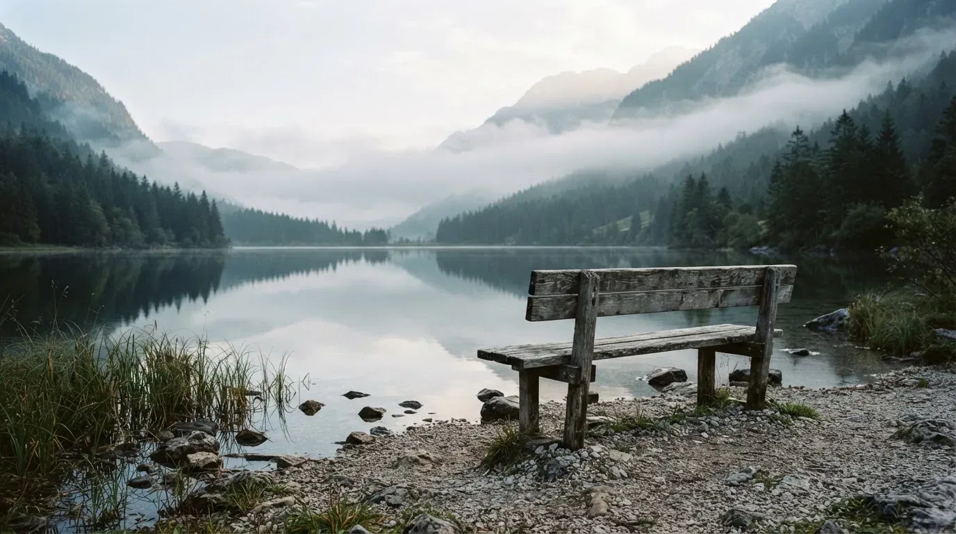 Eine Holzbank am Ufer eines ruhigen Bergsees, am gegenüberliegenden Ufer steigen dunkle Hänge in den Frühnebel