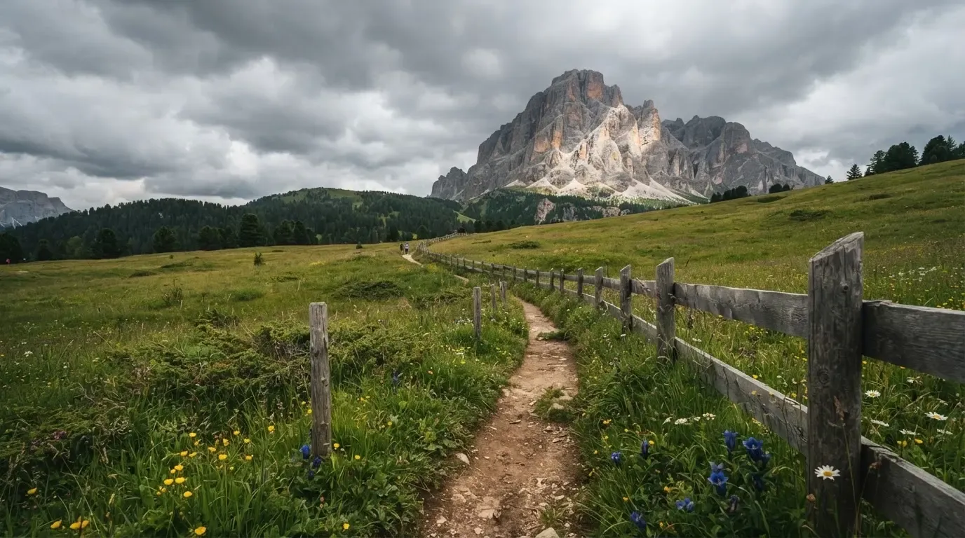 Ein Feldweg führt durch eine Alpwiese mit Holzzaun, im Hintergrund hängen Wolken über einem felsigen Gipfel