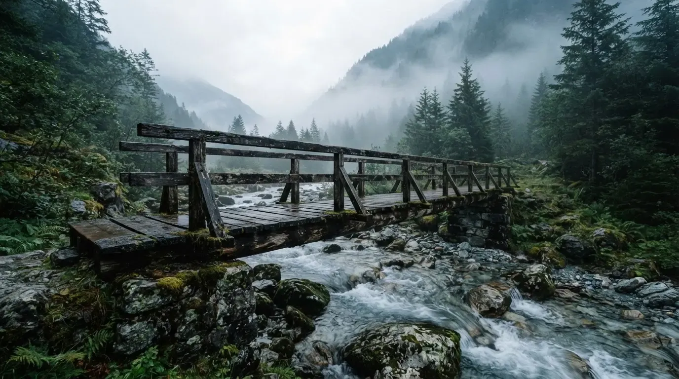 Eine alte Holzbrücke über einen Gebirgsbach, Moos auf den Steinen, Nebelschwaden hängen im Tal