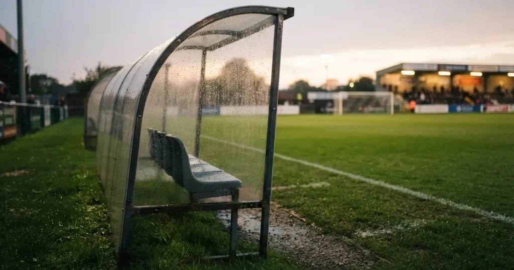 Eine leere Ersatzbank unter einem Plexiglasdach am Spielfeldrand, Regentropfen auf der Scheibe, dahinter ein Rasenplatz
