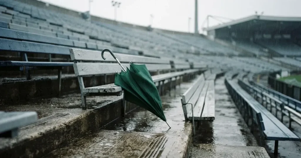 Ein offener Regenschirm lehnt an einer leeren Tribünenbank, Regen fällt auf die Stufen eines Stadions