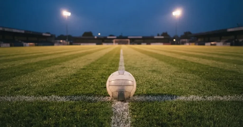Ein Fussball liegt auf dem Anstosspunkt eines leeren Rasenplatzes in der Abenddämmerung, im Hintergrund leuchten Flutlichtmasten.