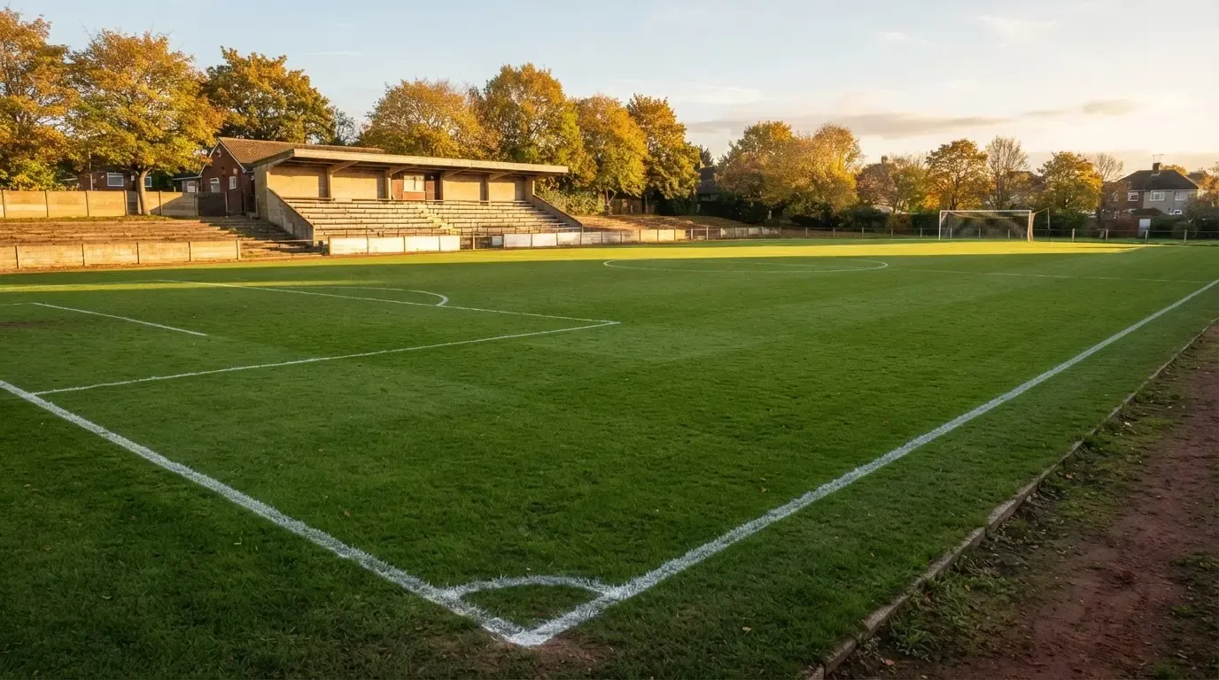 Leere Zuschauerränge eines kleinen Stadions im Herbstlicht, davor ein Rasenplatz mit frisch gezogenen Linien.