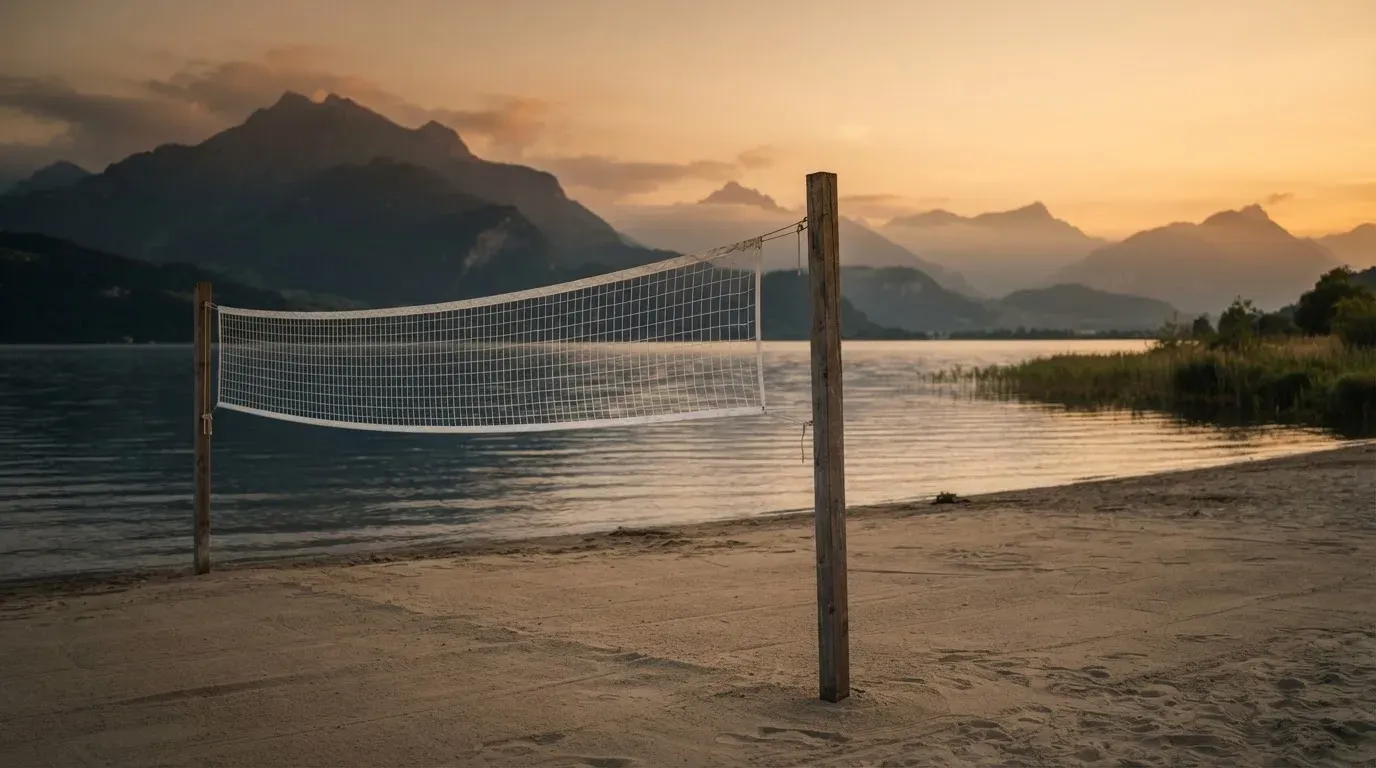 Ein Volleyballnetz steht an einem Seeufer in der Abenddämmerung, der Sand ist leer und das Wasser glatt.