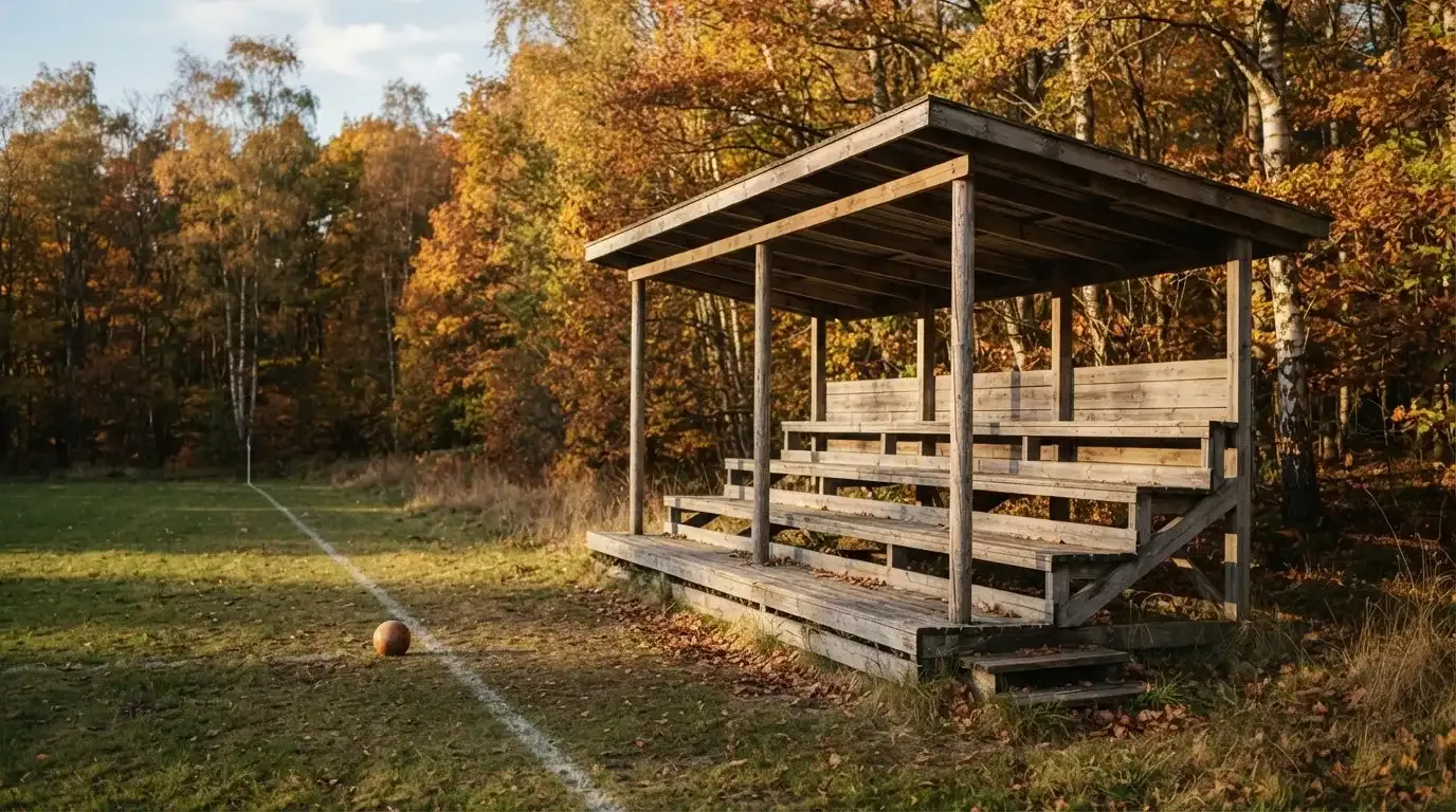 Eine leere Holztribüne steht am Waldrand neben einem kleinen Sportplatz im Herbstlicht.