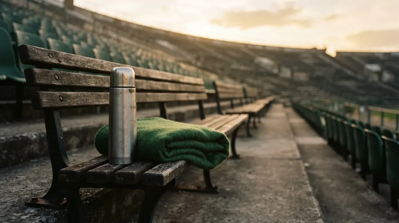 Eine Thermoskanne und eine zusammengelegte Wolldecke stehen auf einer leeren Stadionbank, die Sonne geht hinter den Rängen unter.